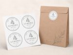 Üzerinde botanik desen bulunan 4 cm çapında Özel Üretim Kuşe etiketin yer aldığı kahverengi bir kağıt torba, üzerinde "Pure. Natural. Care. Est. 2024" yazan ve sade bir yaprak motifine sahip dört adet uyumlu Lumora etiketinin bulunduğu bir sayfanın yanında duruyor.
